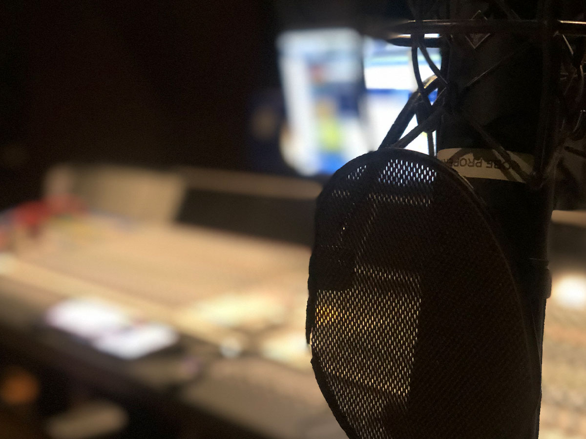 Close up of a microphone in a recording studio with a blurry sound board in the background.